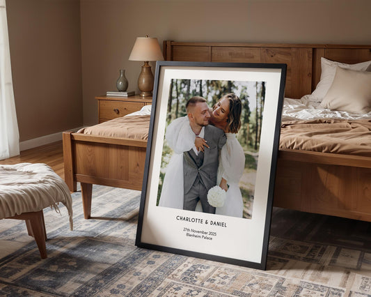 A personalised wedding photo print featuring a couple on their wedding day in elegant natural light. The bride wears an off-shoulder white gown and holds a soft green and white bouquet while the groom embraces her. Minimal modern layout with names, wedding date and venue below the image. A stylish custom keepsake for couples, weddings, anniversaries or home decor.